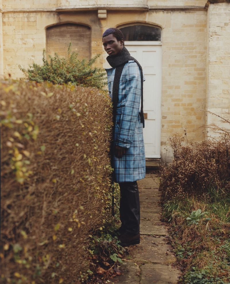 Dark-skinned male model in blue plaid coat stands outside a beige brick building, posing next to a trimmed hedge, fashion editorial.