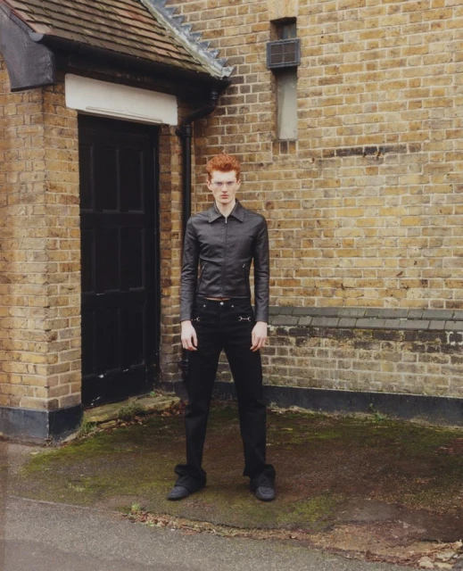 Red-haired man in black jacket and jeans stands in front of a brick building, posing in an outdoor urban setting.
