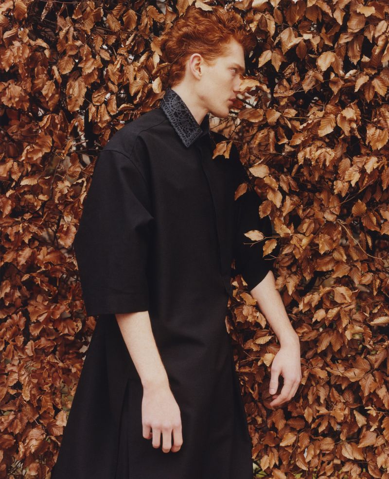A male fashion model with red hair stands against a backdrop of dry autumn leaves wearing a black shirt and skirt ensemble.