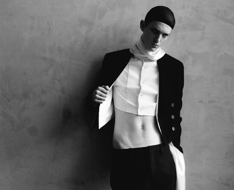 Monochromatic portrait of a slender adult male model in a black blazer and white shirt, standing against a textured gray wall.