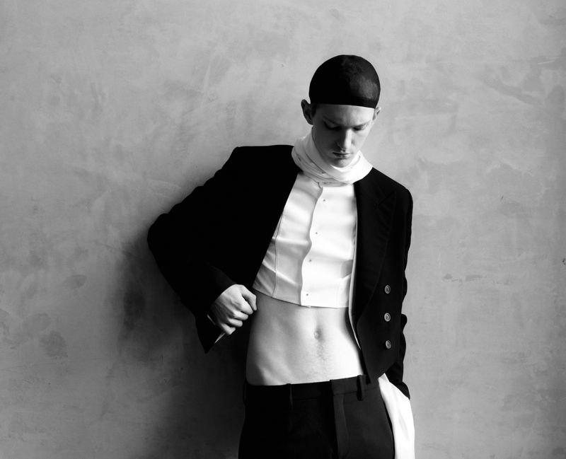 Monochrome portrait of a shirtless man in a cropped blazer with a scarf, looking down, in a minimalist studio setting with textured wall.