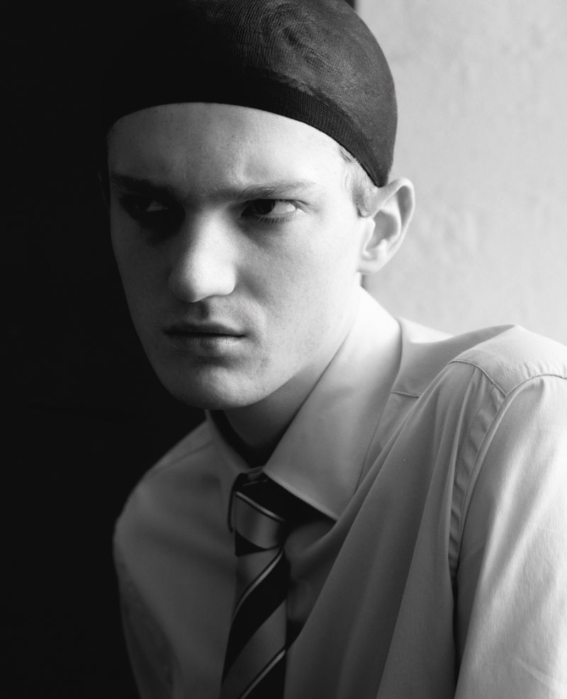 Monochrome portrait of a fair-skinned adult male wearing a cap, white shirt, and striped tie against a stark, contrasting background.