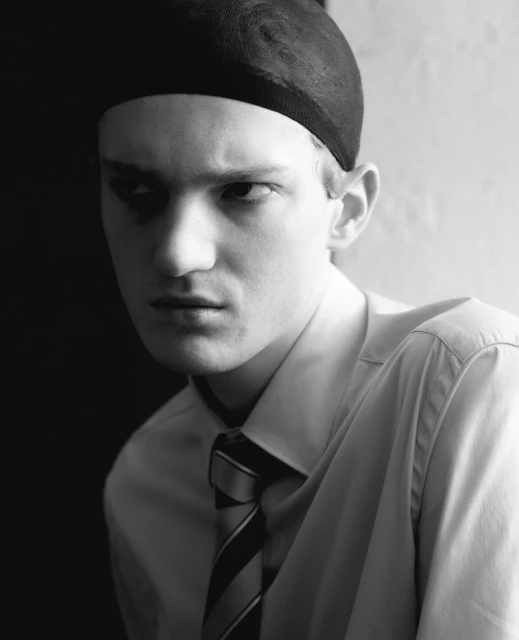 Monochrome portrait of a fair-skinned adult male wearing a cap, white shirt, and striped tie against a stark, contrasting background.