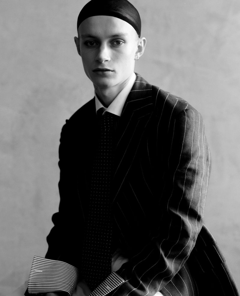 Monochrome portrait of a fair-skinned adult male wearing a pinstripe suit, tie, and black cap, set against a neutral, textured backdrop.
