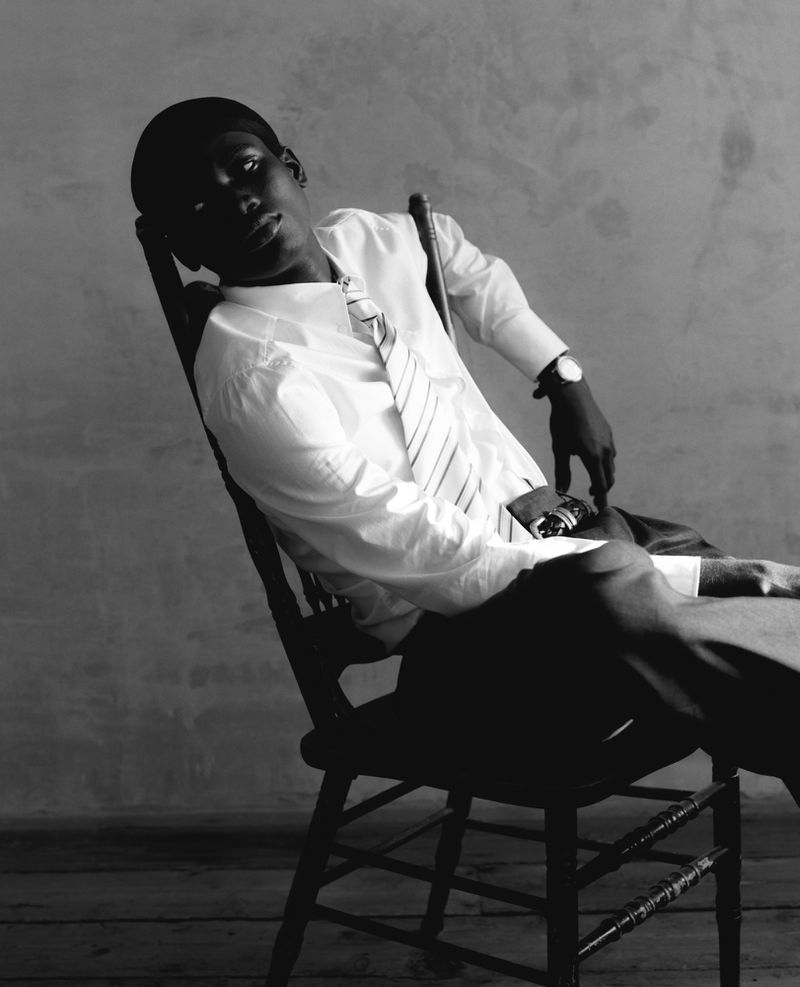 Black and white portrait of a dark-skinned adult male in dress shirt and tie, leaning back in a chair against a textured background.