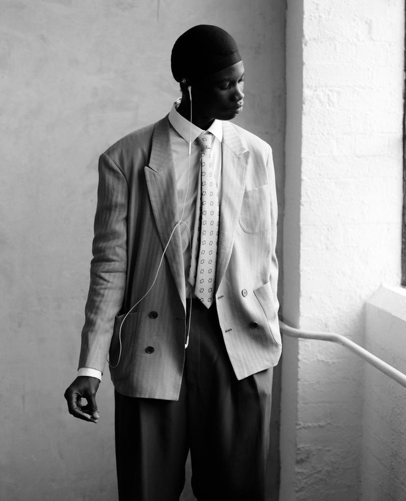 Black man in an oversized grey suit with earphones standing indoors near a window, bathed in soft natural light in black and white.