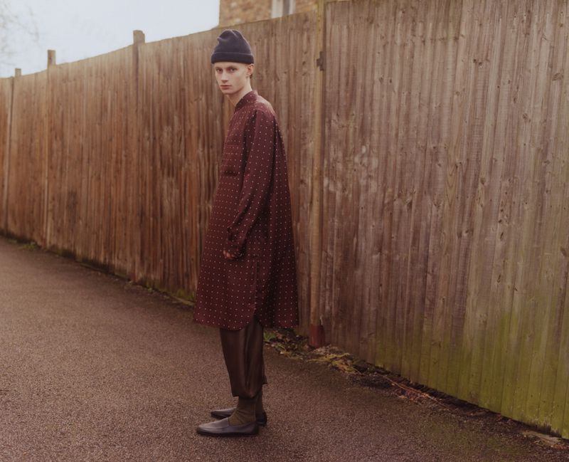 A young adult male stands against a wooden fence in a long-sleeved dotted shirt, dark trousers, and a beanie, outdoors on an overcast day.