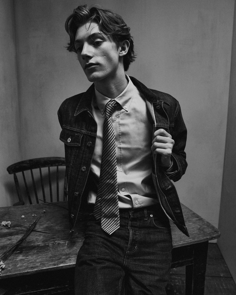 Monochrome portrait of a young adult male in denim jacket and striped tie, leaning against a wooden table in a studio setting.