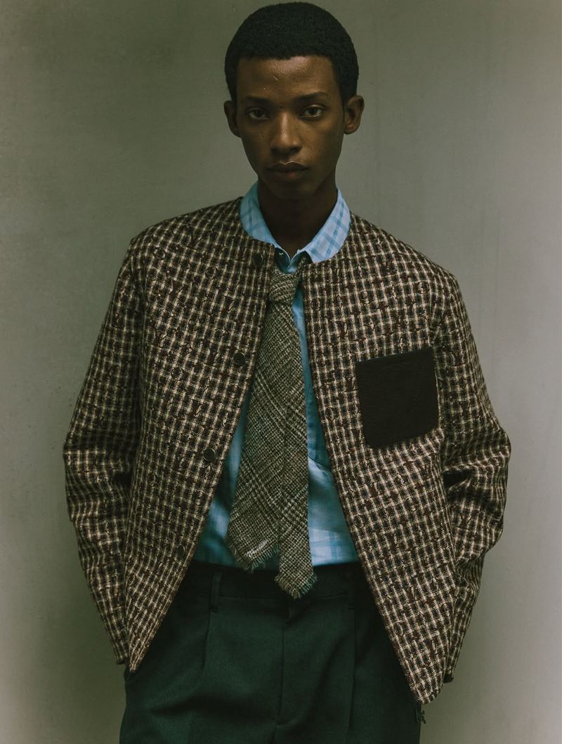 Adult male in a brown and white checkered jacket, light blue shirt, and green pants, stands against a neutral wall in a studio setting.