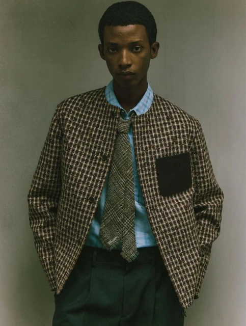 Adult male in a brown and white checkered jacket, light blue shirt, and green pants, stands against a neutral wall in a studio setting.