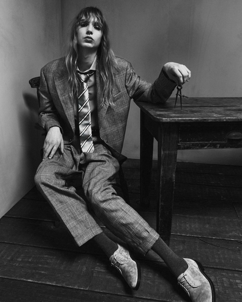 Monochrome portrait of a young adult with long hair in a suit and tie, seated at a wooden table, holding a drawing compass.