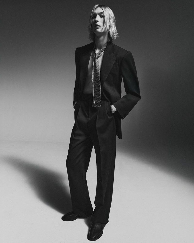 Monochrome portrait of a male fashion model wearing a suit and tie, posing in a photo studio with soft lighting and neutral background.