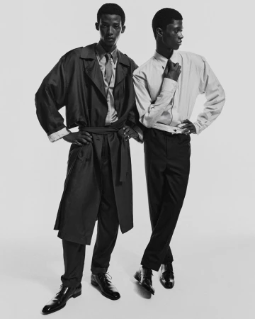 Two Black men in stylish attire, one in a trench coat, the other in a shirt and tie, both in dress pants and shoes, posing in a studio.
