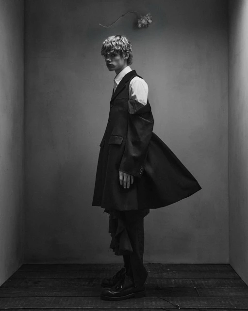 Young man in dark, oversized blazer and suit pants poses in studio with gray walls and wooden floor, under a suspended white flower.