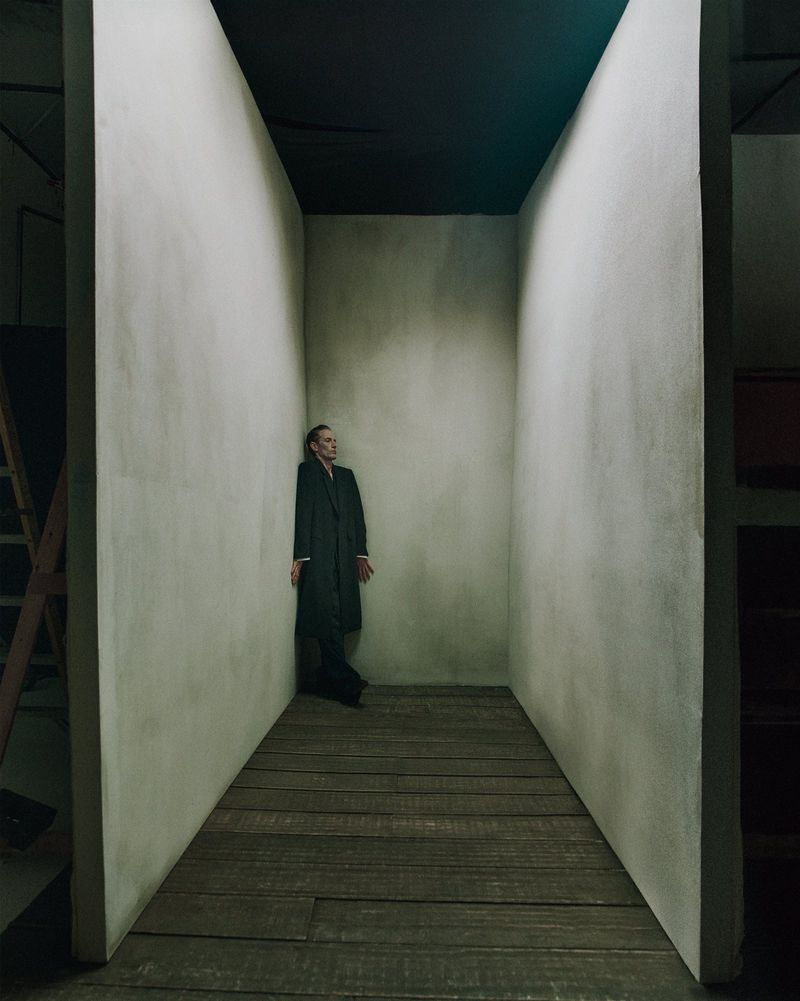 Adult male in a dark coat stands in a narrow hallway with wooden floors and white walls under dim light, creating a contemplative scene.