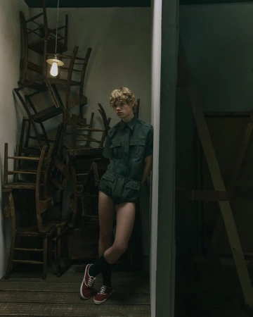 Person with blond hair wears a short olive green playsuit and red sneakers in front of a pile of wooden chairs indoors.