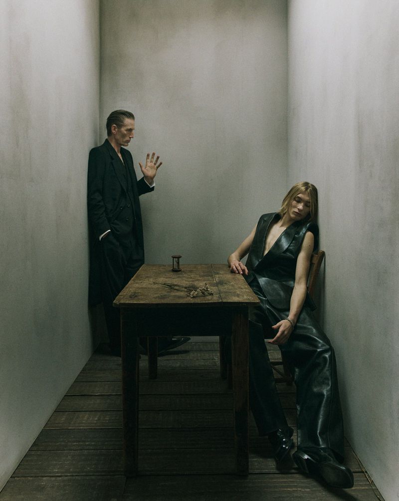 Fashion shot of two people in a stark room, one man standing with his hand raised and the other woman sitting at a table with an hourglass.
