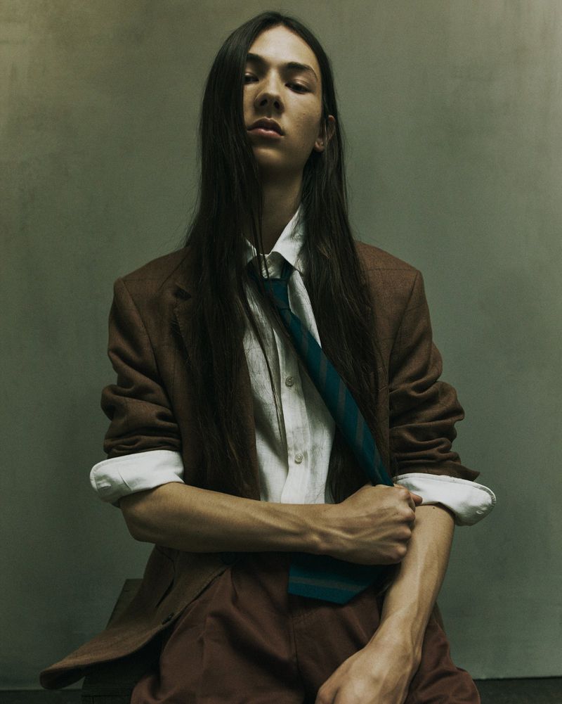 Fashionable young adult posing indoors wearing a brown blazer, white shirt, and tie in a portrait with muted, neutral tones.