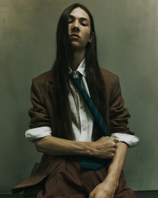 Fashionable young adult posing indoors wearing a brown blazer, white shirt, and tie in a portrait with muted, neutral tones.