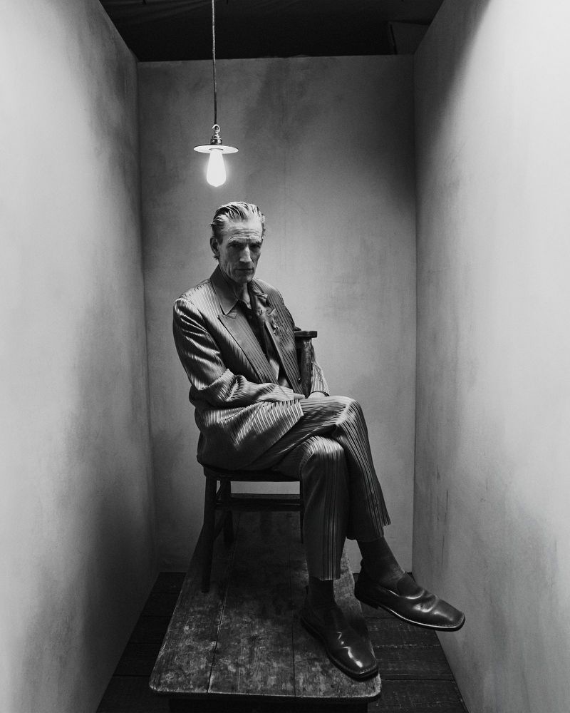 Older man in a suit sits on a wooden chair in a minimal gray room under a hanging lightbulb. Black and white portrait, fashion.