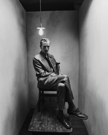 Older man in a suit sits on a wooden chair in a minimal gray room under a hanging lightbulb. Black and white portrait, fashion.