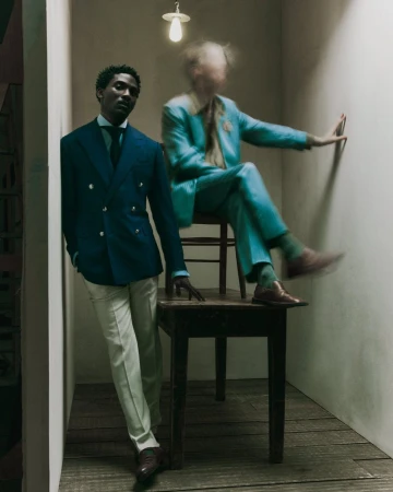 Two men in suits stand indoors: one in a blue suit leaning against a wall, and the other blurred sitting on a table in an aqua suit.