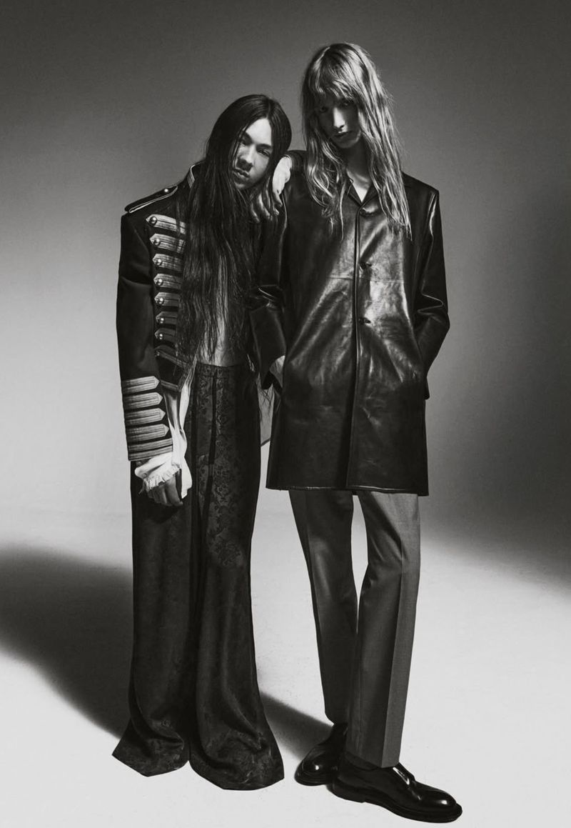 Two people in black and white. One with long hair in black jacket, the other in vintage army-style jacket in a studio setting.
