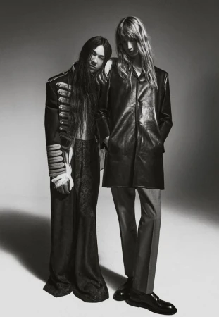 Two people in black and white. One with long hair in black jacket, the other in vintage army-style jacket in a studio setting.