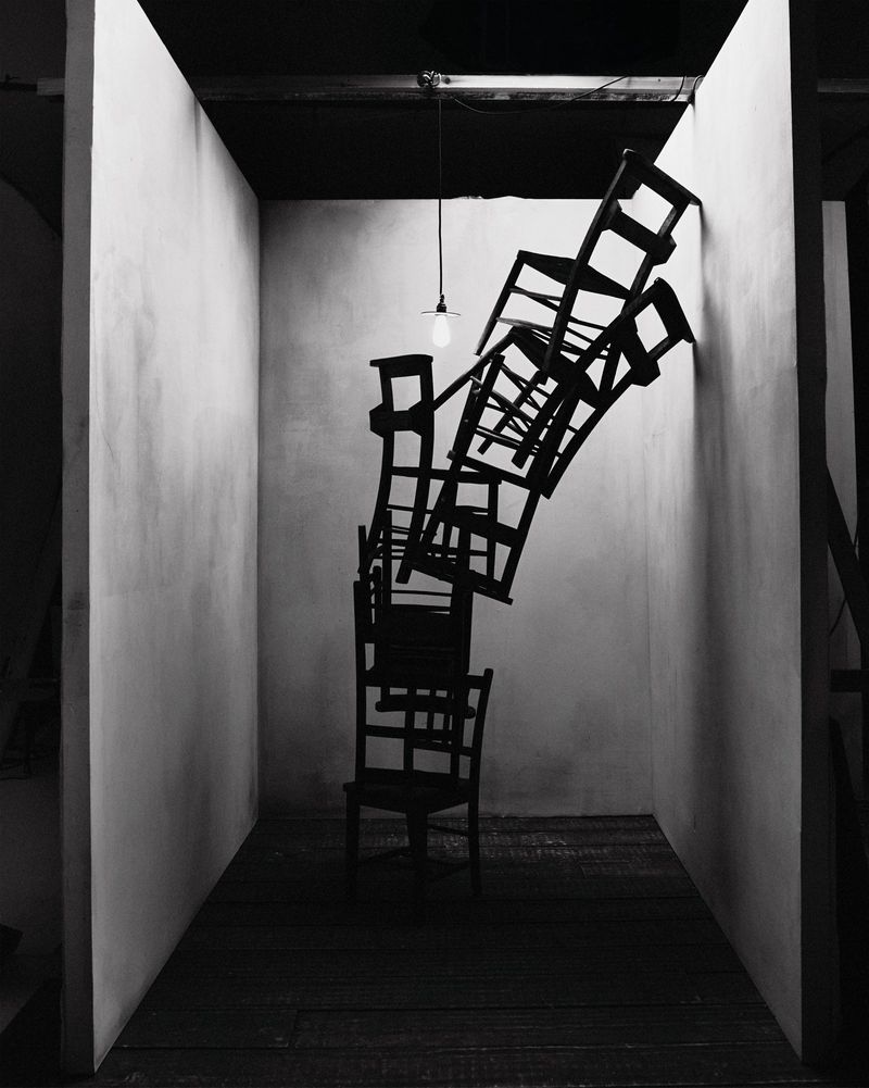 Stack of wooden chairs inside a white walled studio. Dramatic lighting from a single bare bulb illuminates the sculpture.