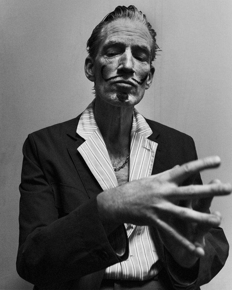 Black-and-white portrait of a mature man with a distinctive mustache and closed eyes, wearing a striped shirt and suit jacket.