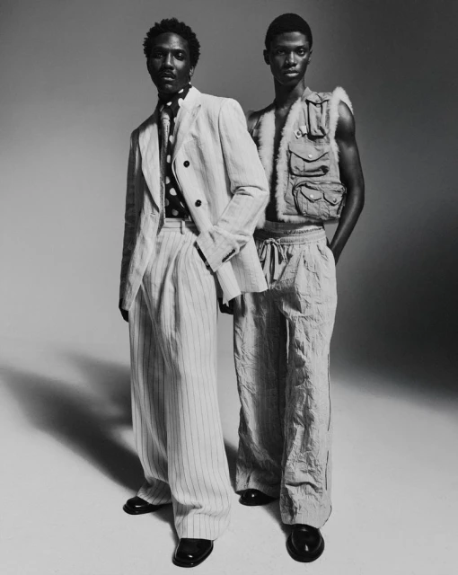 Two Black men in studio fashion shot. Man on left in pinstripe suit with black polka dot shirt, man on right in utility vest and wide pants.