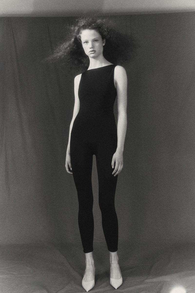 Fashion model poses in black jumpsuit and off-white heels in a studio setting with a neutral gray backdrop, captured in black and white.