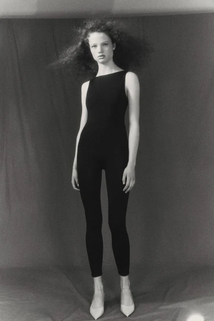 Fashion model poses in black jumpsuit and off-white heels in a studio setting with a neutral gray backdrop, captured in black and white.