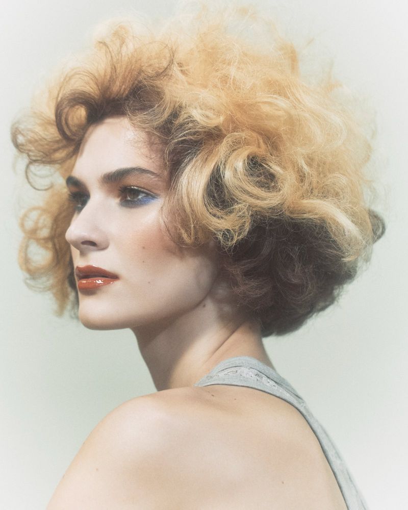 Profile of a young adult woman with voluminous blonde hair, wearing a gray tank top, and striking makeup against a soft background.