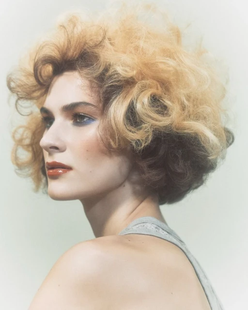 Profile of a young adult woman with voluminous blonde hair, wearing a gray tank top, and striking makeup against a soft background.