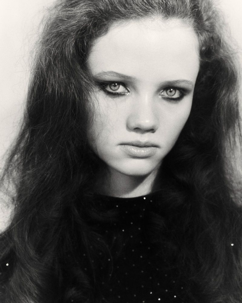 Monochrome portrait of a young woman with long, dark wavy hair and striking eye makeup looking directly at the camera. Neutral background.
