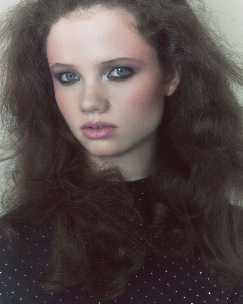 Portrait of a young woman with curly brown hair and grey eyes, wearing a black top with dots against a neutral background.