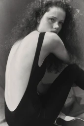 Model with long curly hair wearing a black backless leotard sitting against a neutral background.