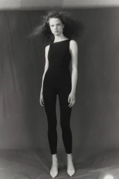 Model in black jumpsuit with voluminous hair standing against a dark seamless studio backdrop