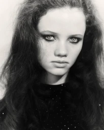 Close-up portrait of a young woman with dark voluminous hair and dark eye makeup wearing a dark top against a light background