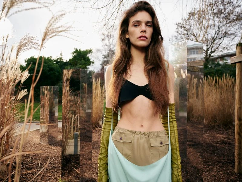 Woman in black top and light green bottoms standing outdoors among tall grass and mirrored columns in an editorial fashion setting.