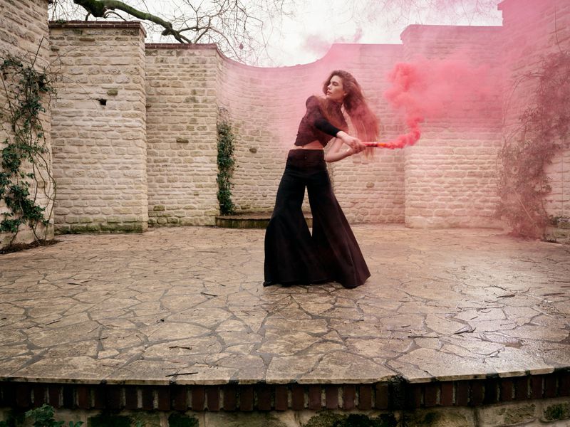 A woman in wide-leg pants poses with a red smoke flare in a walled courtyard with stone pavers and a stone wall covered with vines.