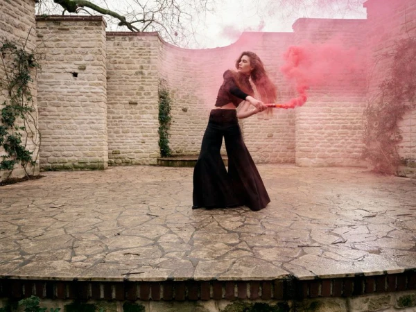 A woman in wide-leg pants poses with a red smoke flare in a walled courtyard with stone pavers and a stone wall covered with vines.