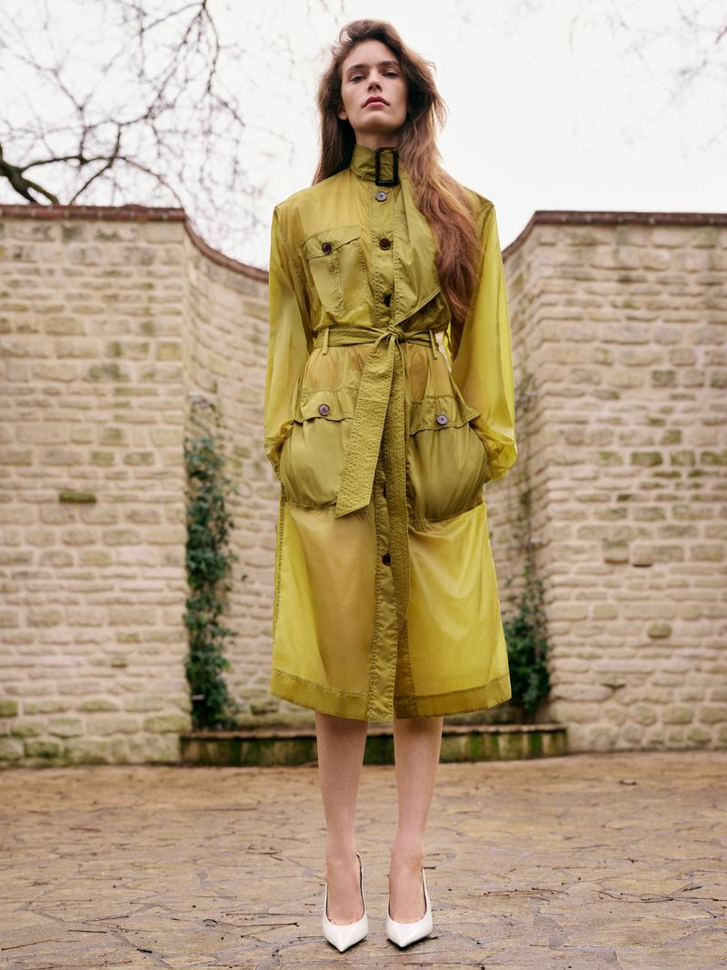 Woman poses in a transparent yellow trench coat with white pointed-toe shoes in an outdoor courtyard with stone walls and pavement.
