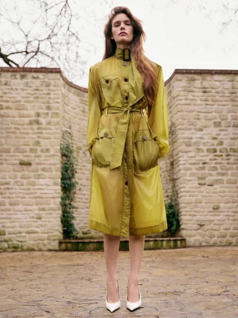 Woman poses in a transparent yellow trench coat with white pointed-toe shoes in an outdoor courtyard with stone walls and pavement.