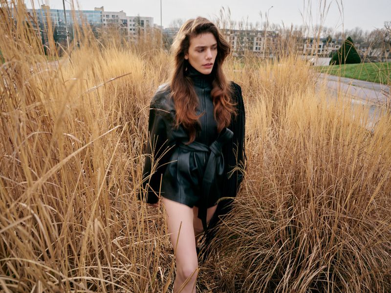 Adult woman in a black belted jacket stands in a field of tall golden grass with buildings in the background on an overcast day.