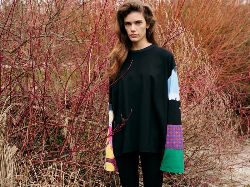 A woman in a black shirt with patterned sleeves stands in front of red-twig dogwood, creating a natural outdoor fashion shot.