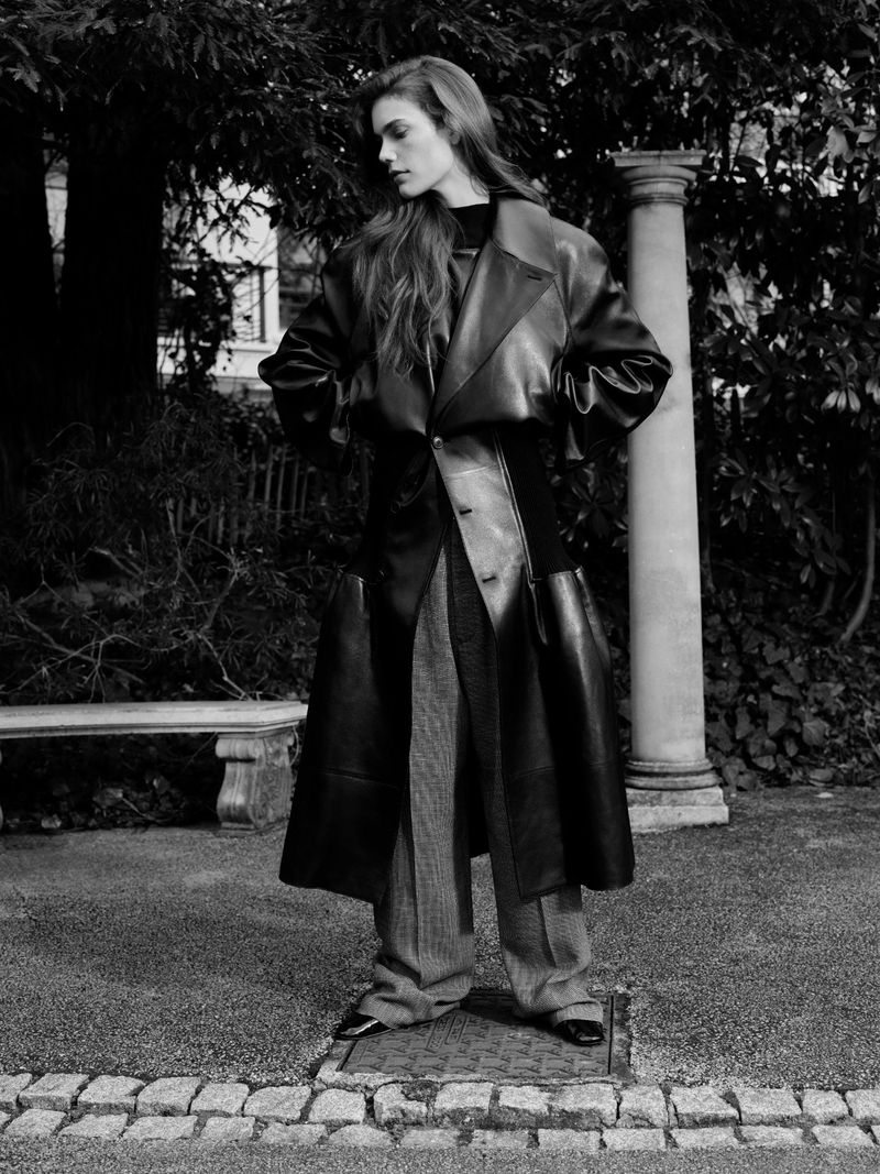 A black and white full shot of a woman with long hair outdoors, wearing a long black leather coat, and grey trousers, with trees in the background.