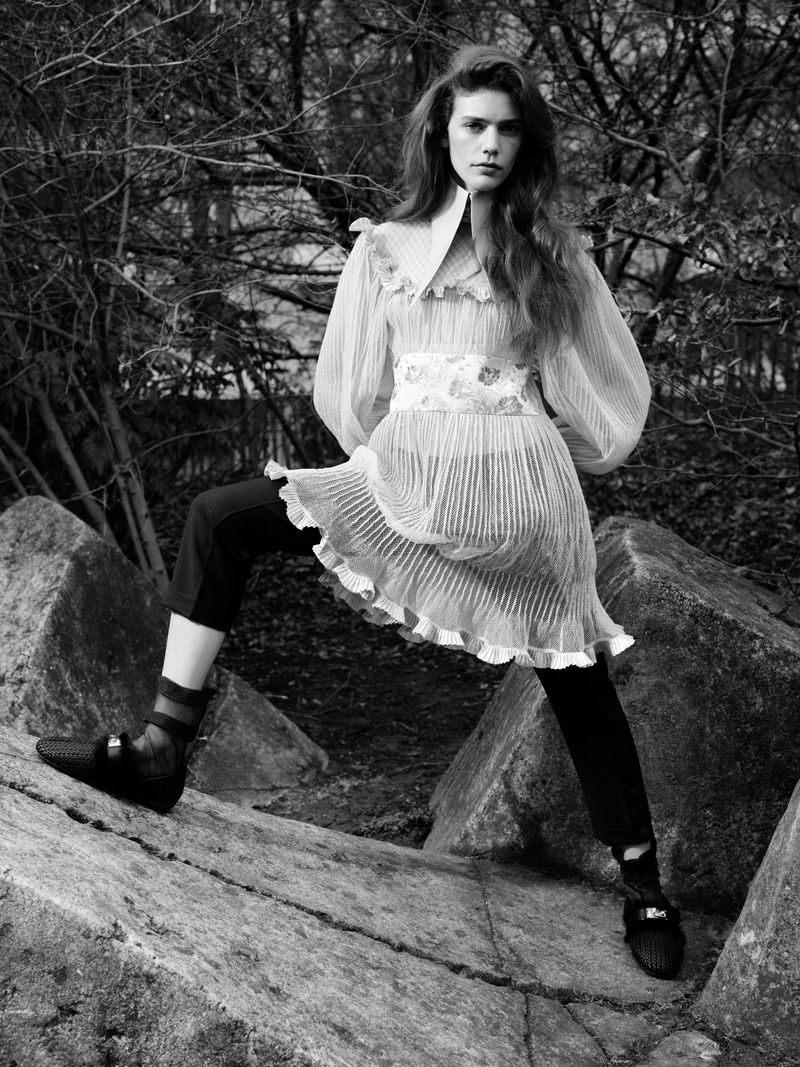A woman stands with one foot on a large rock, wearing a sheer pleated mini dress with a wide waistband and dark trousers in a wooded area.