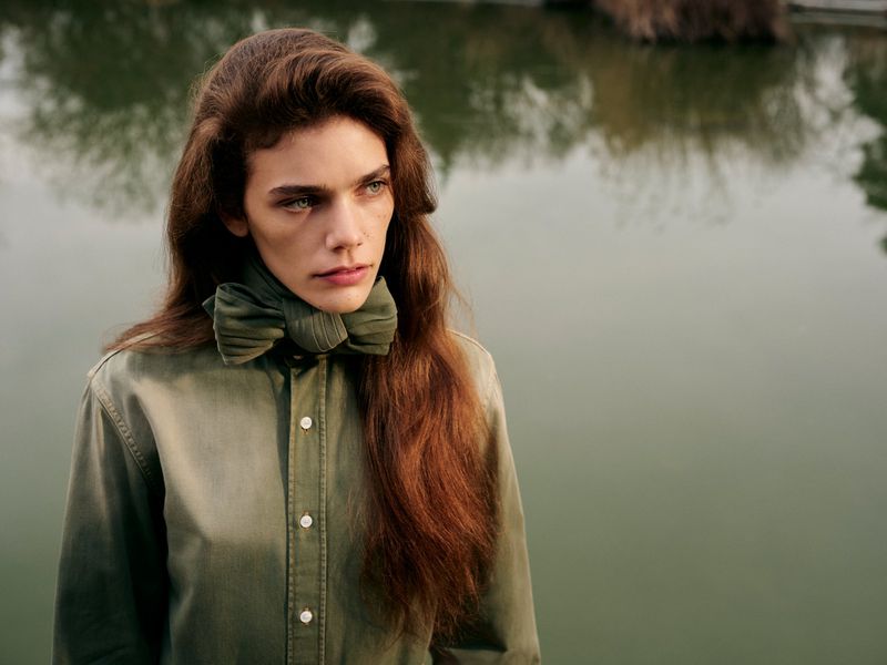 A model with long auburn hair poses near a lake wearing a green collared shirt and matching neck piece in a medium shot outdoors.
